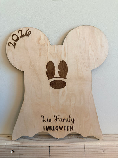 A personalized wooden ghost-shaped plaque with a Mickey Mouse silhouette, featuring the engraved text '2026' at the top left and 'Lin Family HALLOWEEN' at the bottom. The plaque is displayed on a wooden shelf against a light gray wall.