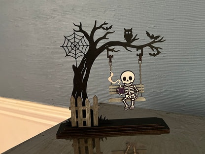 Handmade laser-cut Halloween skeleton décor – spooky skeleton sipping coffee on a swing. Freestanding, hand painted & customizable.

