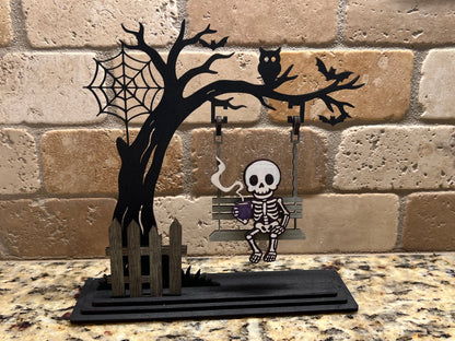 Handmade laser-cut Halloween skeleton décor – spooky skeleton sipping coffee on a swing. Freestanding, hand painted & customizable.
